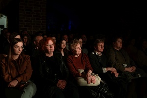 Koncert: Pracownia piosenki prezentuje - Niepozorny zaścianek - Zdjęcie pokazuje widownię w Piwnicach TCK podczas koncertu. Widzowie, siedzący na krzesłach obserwują występ. Na zdjęciu widać różnorodność publiczności, od młodych po starszych słuchaczy. Sala jest delikatnie oświetlona.