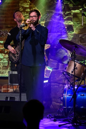 Koncert jazzowy: Tomasz Chyła Quintet - Zdjęcie sceny z koncertu Tomasz Chyła Quintet, pokazujące zaangażowanych muzyków. Tomasz Chyła gra na skrzypcach, w tle pozostali muzycy na różnych instrumentach. Scena jest dynamicznie oświetlona, co dodaje energii występowi.