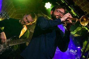 Koncert jazzowy: Tomasz Chyła Quintet - Zdjęcie sceny z koncertu Tomasz Chyła Quintet, pokazujące zaangażowanych muzyków. Tomasz Chyła gra na skrzypcach, w tle pozostali muzycy na różnych instrumentach. Scena jest dynamicznie oświetlona, co dodaje energii występowi.