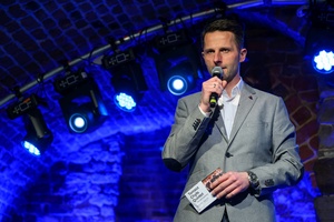 Koncert jazzowy: Tomasz Chyła Quintet - Konferansjer Sebastian Szafarz wita publiczność i zapowiada koncert Tomasz Chyła Quintet, stoi na oświetlonej scenie.