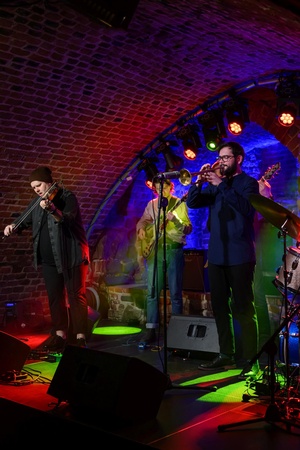 Koncert jazzowy: Tomasz Chyła Quintet - Zdjęcie sceny z koncertu Tomasz Chyła Quintet, pokazujące zaangażowanych muzyków. Tomasz Chyła gra na skrzypcach, w tle pozostali muzycy na różnych instrumentach. Scena jest dynamicznie oświetlona, co dodaje energii występowi.